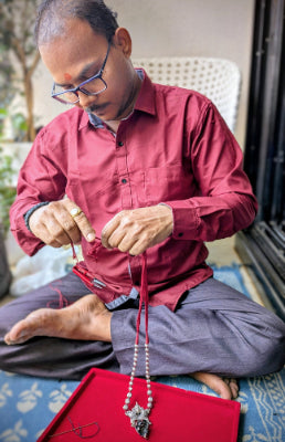 Man sitting on a mat, wearing a red shirt and gray pants, working on a red bag with beads.